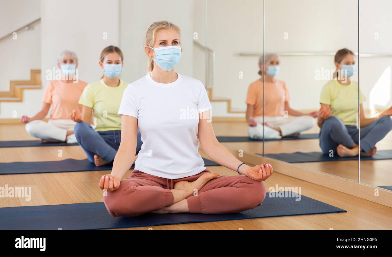 Woman in mask sitting in lotus pose while training yoga with her family