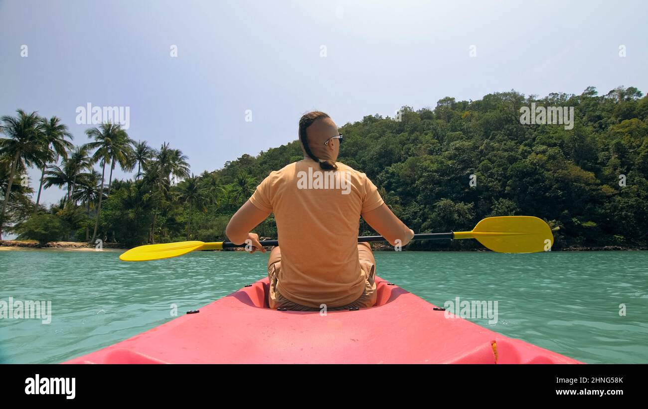 Young man with sunglasses and hat rows pink plastic canoe along sea against green hilly islands with wild jungles. Traveling to tropical countries. Strong guy is sailing on kayak in ocean, back view. Stock Photo