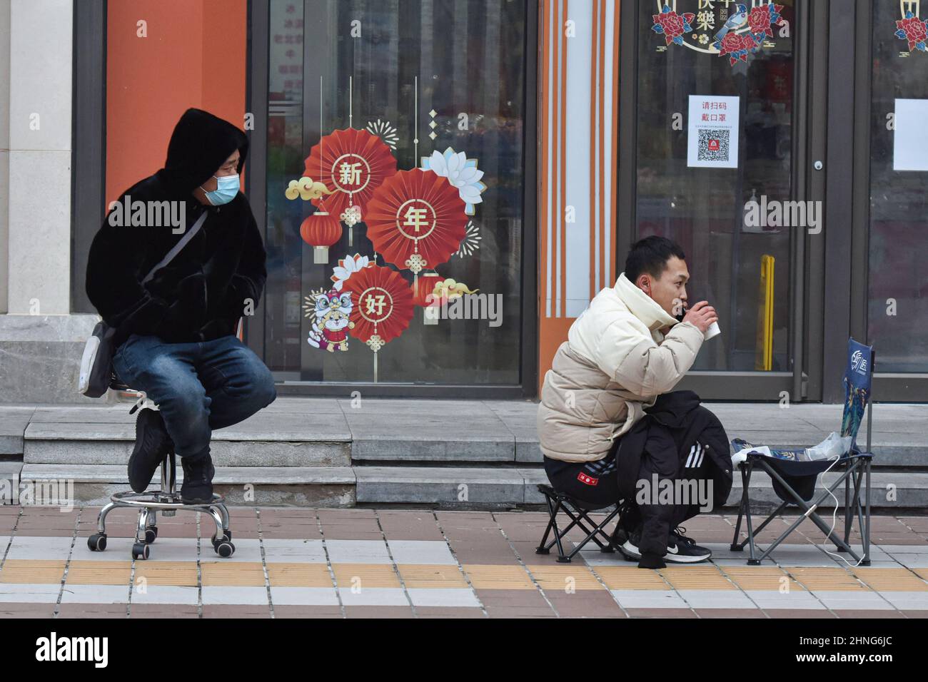 A customer sitting on low chair takes hot water while queuing up to buy 'Bing Dwen Dwen'. Every day since the opening of the Beijing Winter Olympics, people have lined up outside the licensed store on Beijing's Wangfujing Street to buy toys featuring the games' mascot, Bing Dwen Dwen, which have become a sought-after item in China. Stock Photo