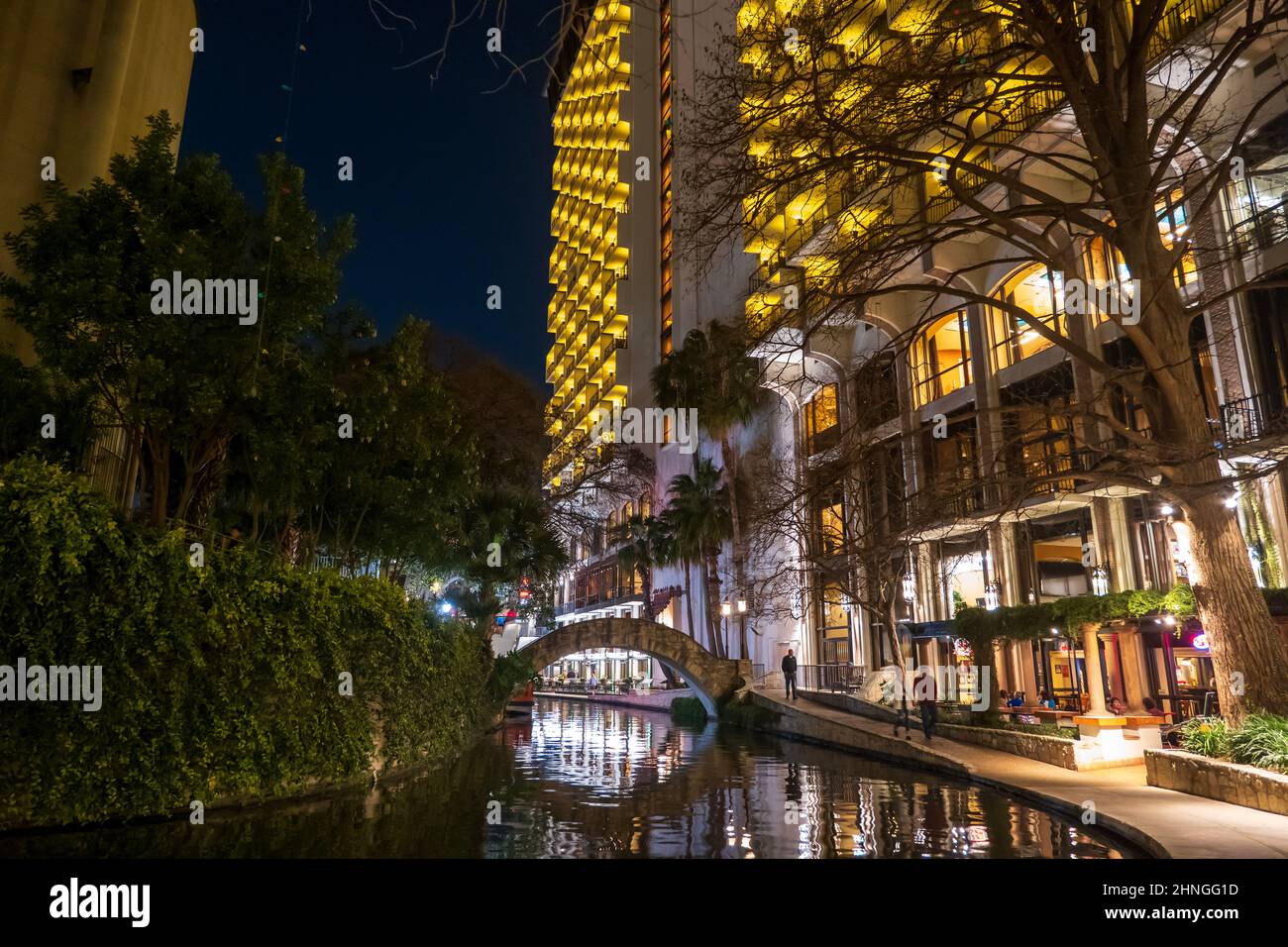 The popular riverwalk in a night scene in downtown San Antonio, Texas, with an arch bridge and tall buildings along the river reflected in the water. Stock Photo