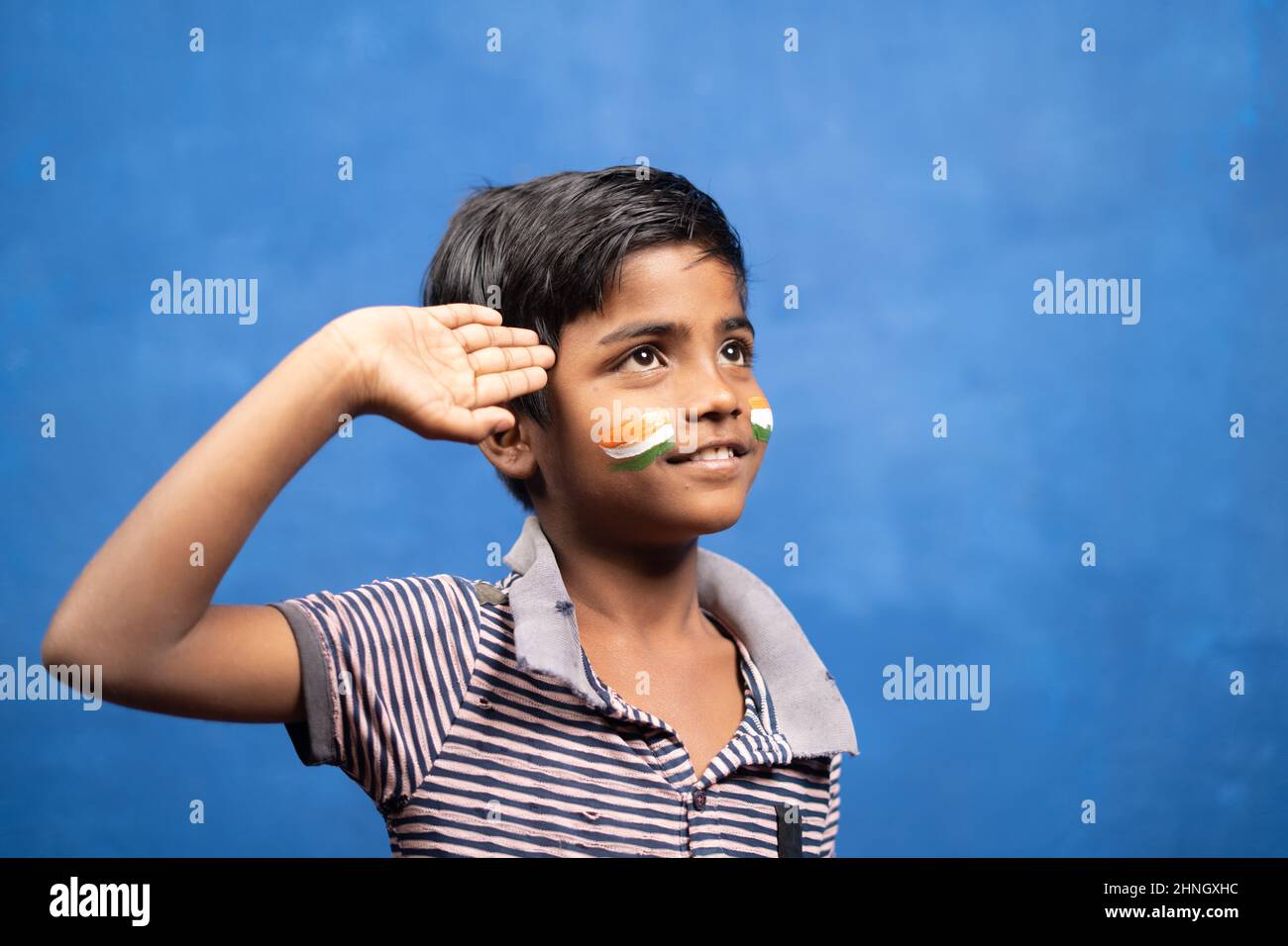 Happy smiling kid with torn shirt and Indian flag on face saluting by looking abovr - concept of poverty, patriotism, freedom and independence day. Stock Photo