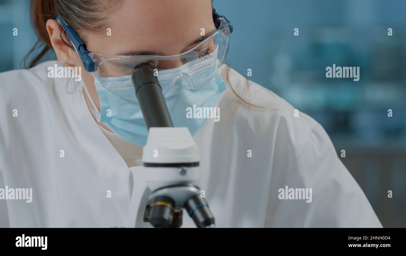 Biologist with face mask using microscope to work on experiment in ...