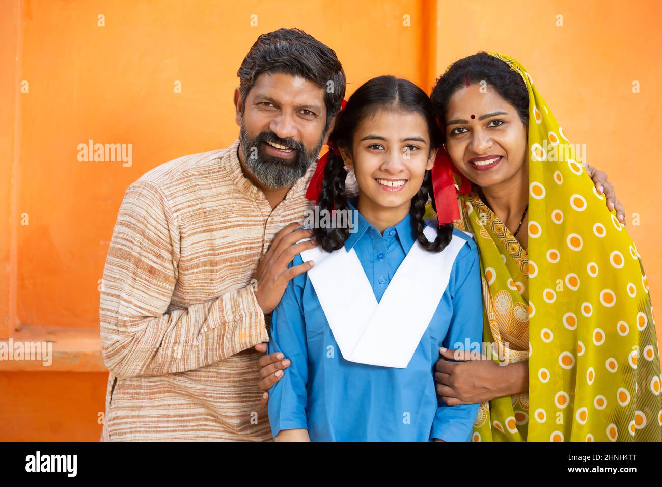 Portrait of happy rural Indian family, young girl in school uniform with her parents, Beard man and woman in sari standing with daughter. looking at c Stock Photo