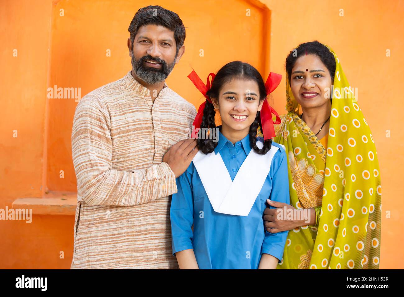 Portrait of happy rural Indian family, young girl in school uniform with her parents, Beard man and woman in sari standing with daughter. looking at c Stock Photo