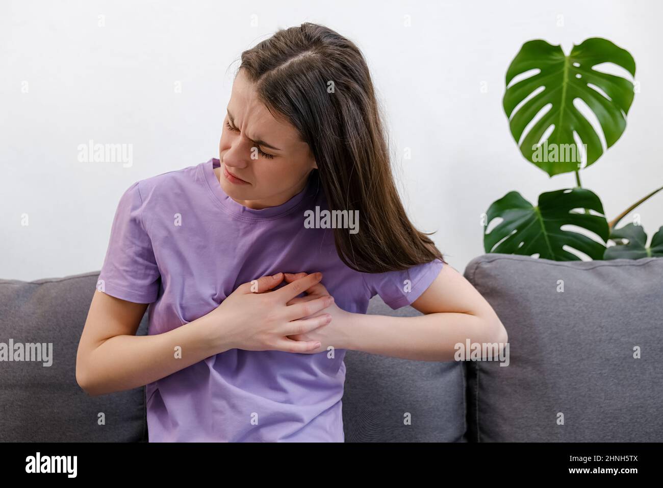 Upset tired young caucasian female feeling pain ache touching chest having heart attack, sad unhappy worried girl suffers from heartache sit on couch Stock Photo