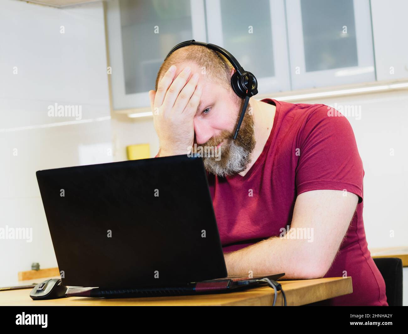 Tired adult person holding head with hand, mature man in headset working from home using laptop and providing online meeting, remote job concept Stock Photo