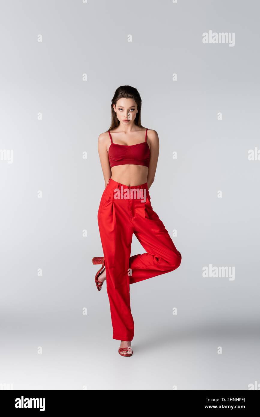 full length of pretty model in red outfit posing while standing on one leg on grey Stock Photo