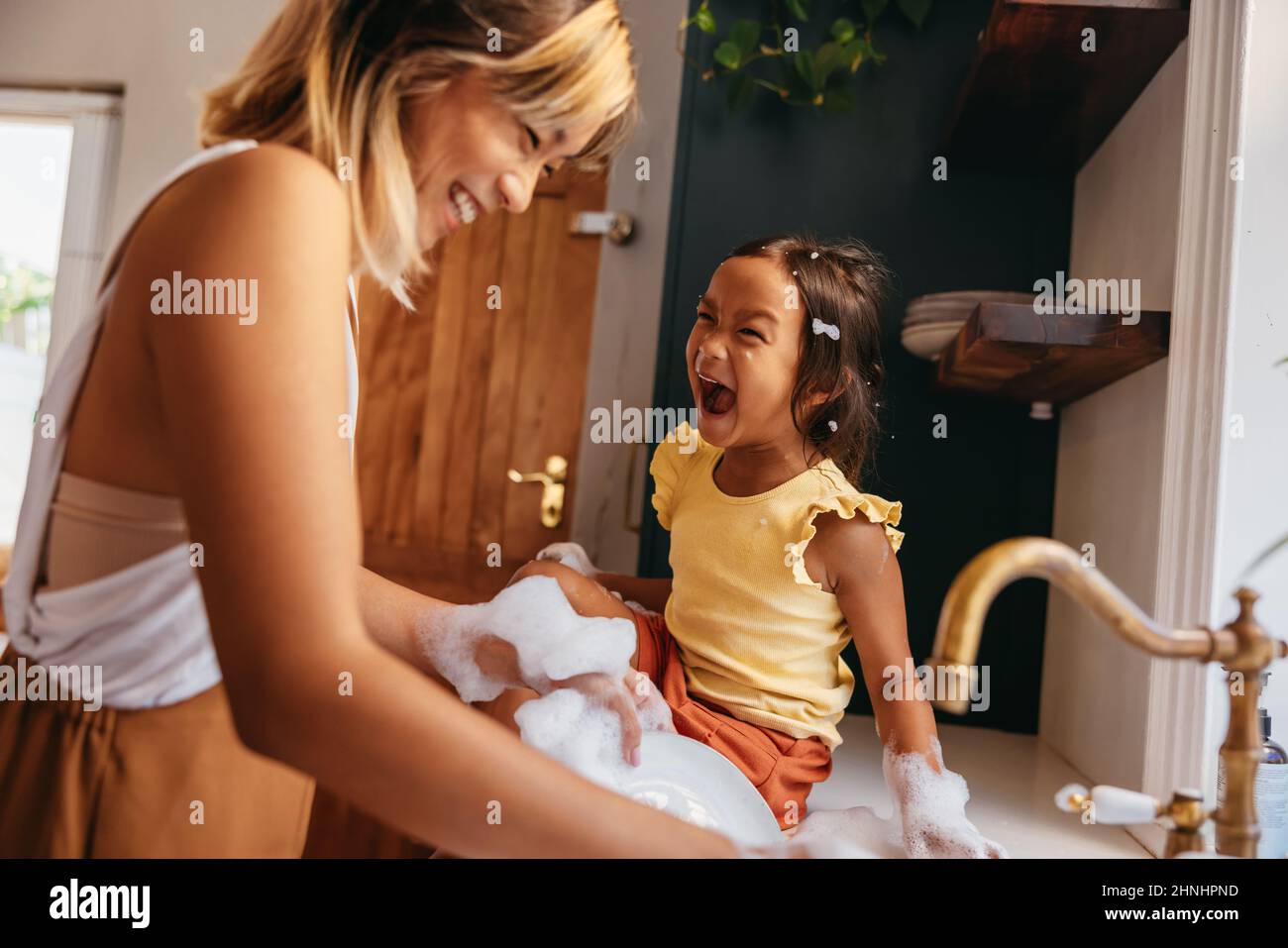 Mother laughing with her daughter while washing the dishes in the kitchen. Cheerful mother and daughter having fun with soap bubbles. Mother and daugh Stock Photo