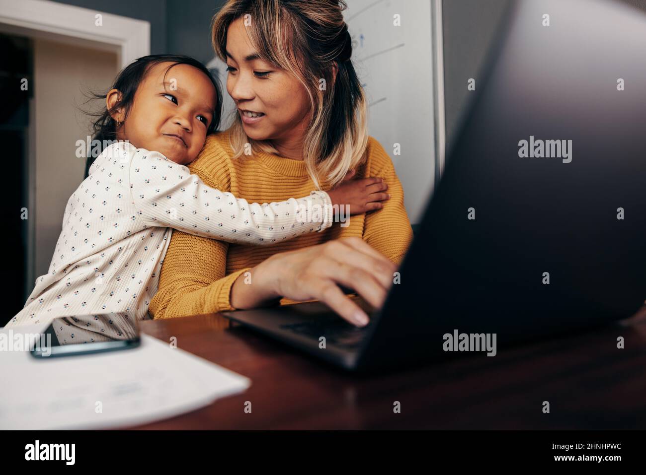 Adorable little girl embracing her mom in her home office. Working mom smiling at her daughter while typing an email on a laptop. Self-employed mother Stock Photo
