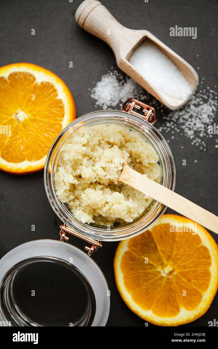 Homemade sugar body scrub in glass jar, decorated with fresh orange slices and wooden spoon with sugar powder on black stone cutting board. Stock Photo