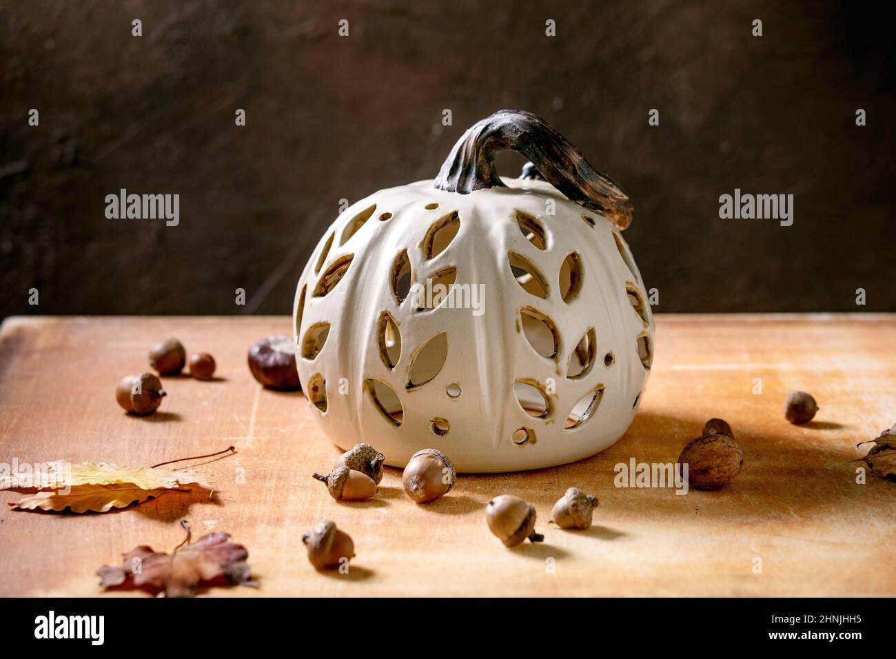 Halloween or Thanksgiving decorations, white handcrafted carved ceramic pumpkin standing on orange stone table with autumn leaves and acorns. Hallowee Stock Photo