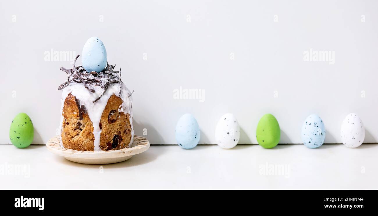 Homemade traditionla Easter kulich cake with chocolate nest, eggs on plate standing on white table with colored eggs in row. Traditional ortodox easte Stock Photo