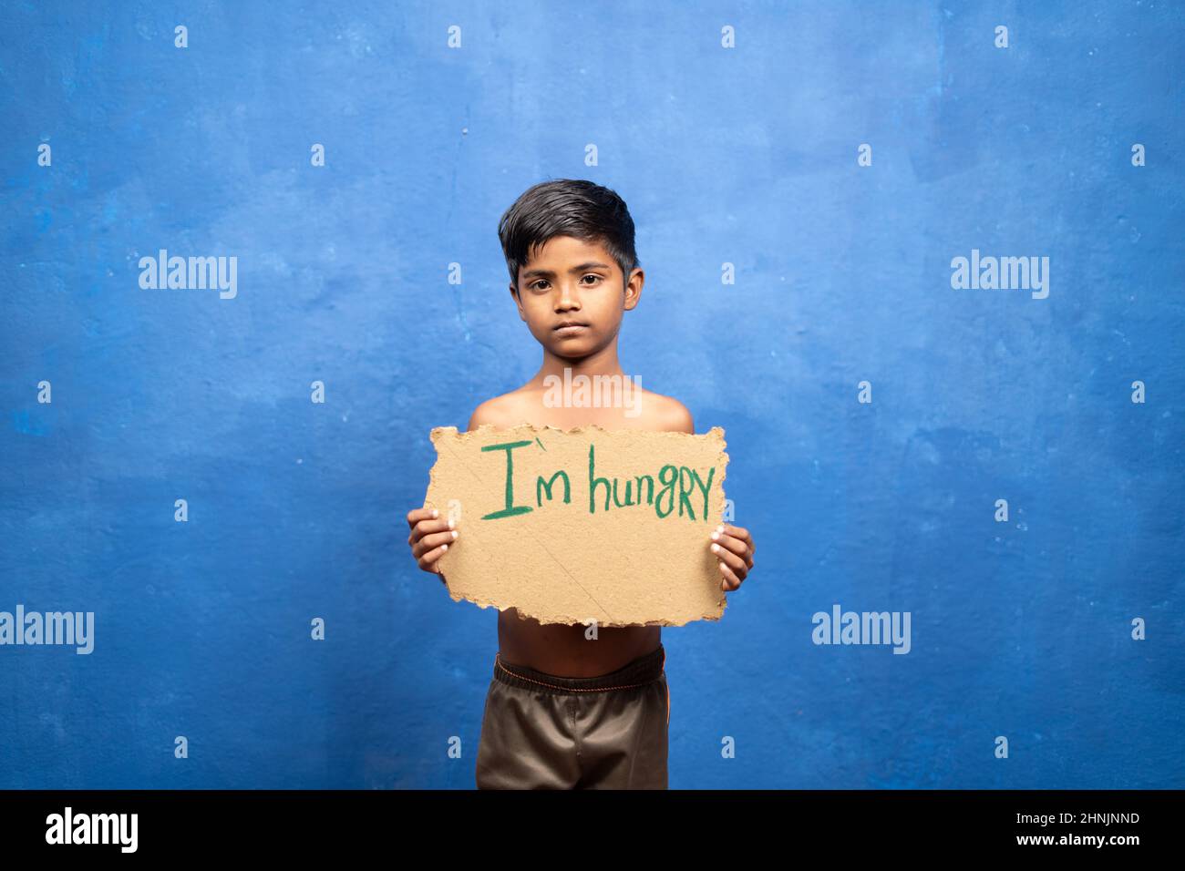 Sad Indian poor kid holding I am hungry sign board by looking at camera on blu background with copy space - concept of poverty and malnutrition. Stock Photo
