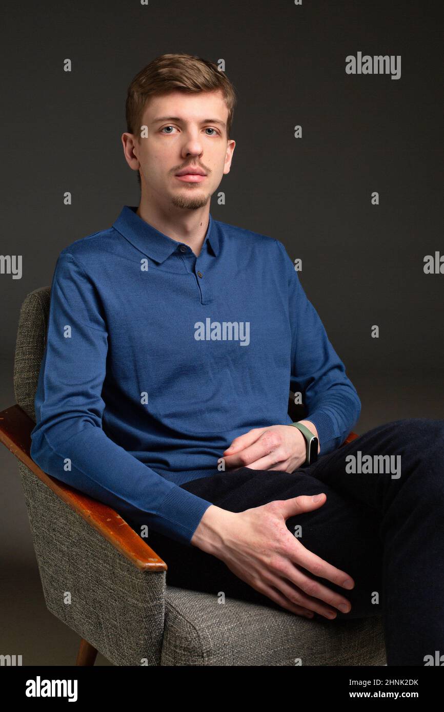 Young blond european man with short beard and mustache isolated on black sitting in armchair Stock Photo