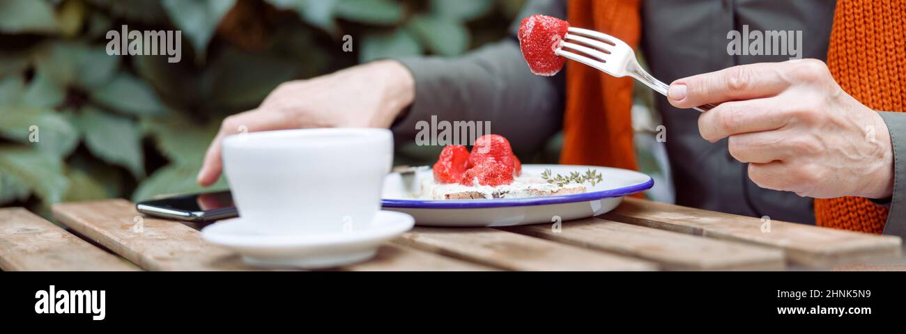 Mature woman eats toast with cream and strawberries taking mobile phone at table on outdoors Stock Photo