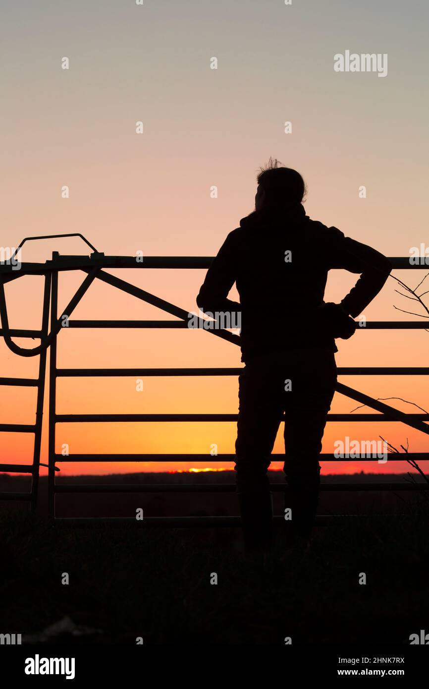 Rear view silhouette of a woman standing isolated outdoors in farmland, by a country gate watching golden, sunset sky. Stock Photo