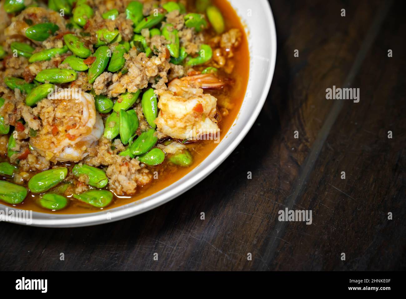 Close up Thai Stir-Fried Stink Beans with Shrimps and spicy shrimp paste sauce in white dish bowl on dark wood table. Stock Photo