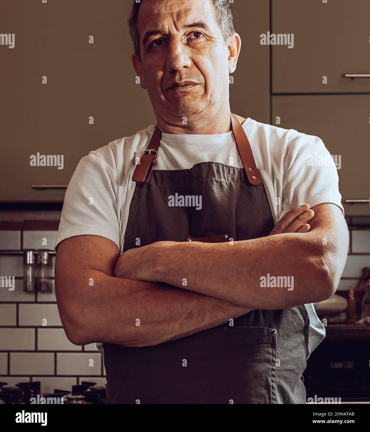 middle-aged cook, muscular in an apron, crossed arms over chest, standing in home kitchen. concept of male householder, chief in home kitchen Stock Photo