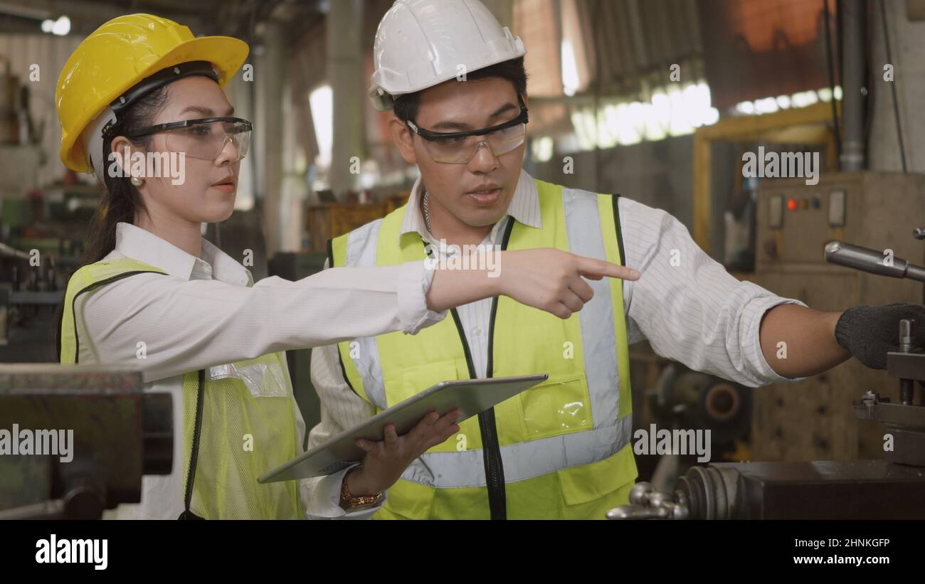 mechanical engineer woman and operation man wearing uniform hardhat and goggles safety working on workshop metal lathe Stock Photo