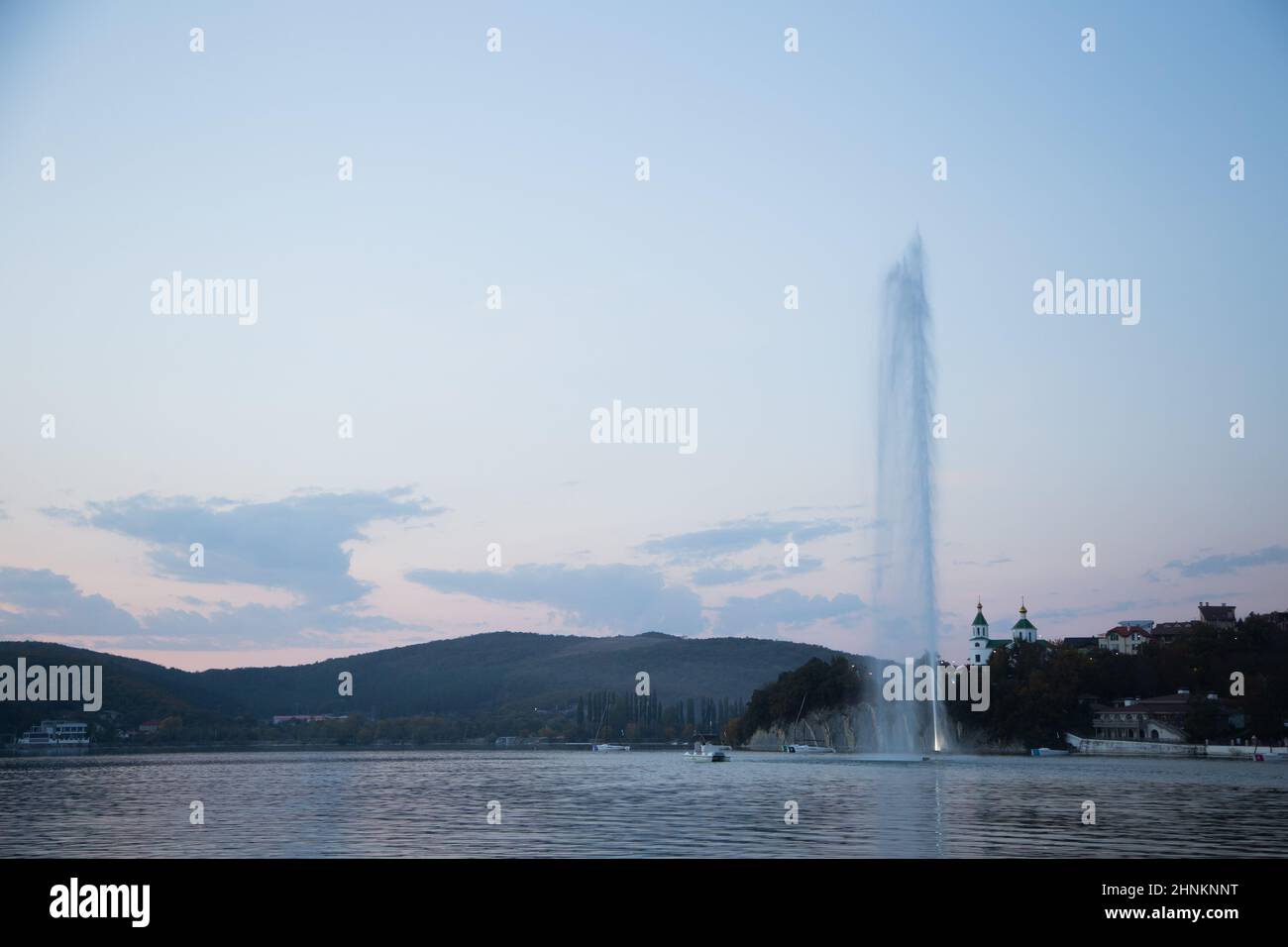 A fountain several meters high spouts from the center of the lake, you can see the mountains and the church, evening. Abrau Durso, Krasnodar Territory. Stock Photo