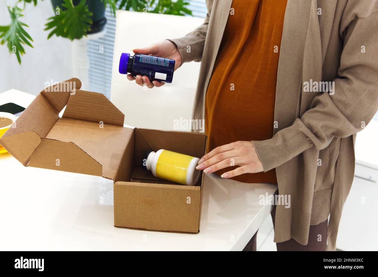 Cropped photo of pregnant woman at home opening cardboard delivery box with bottles of prenatal vitamins and supplements, expectant mother receiving o Stock Photo