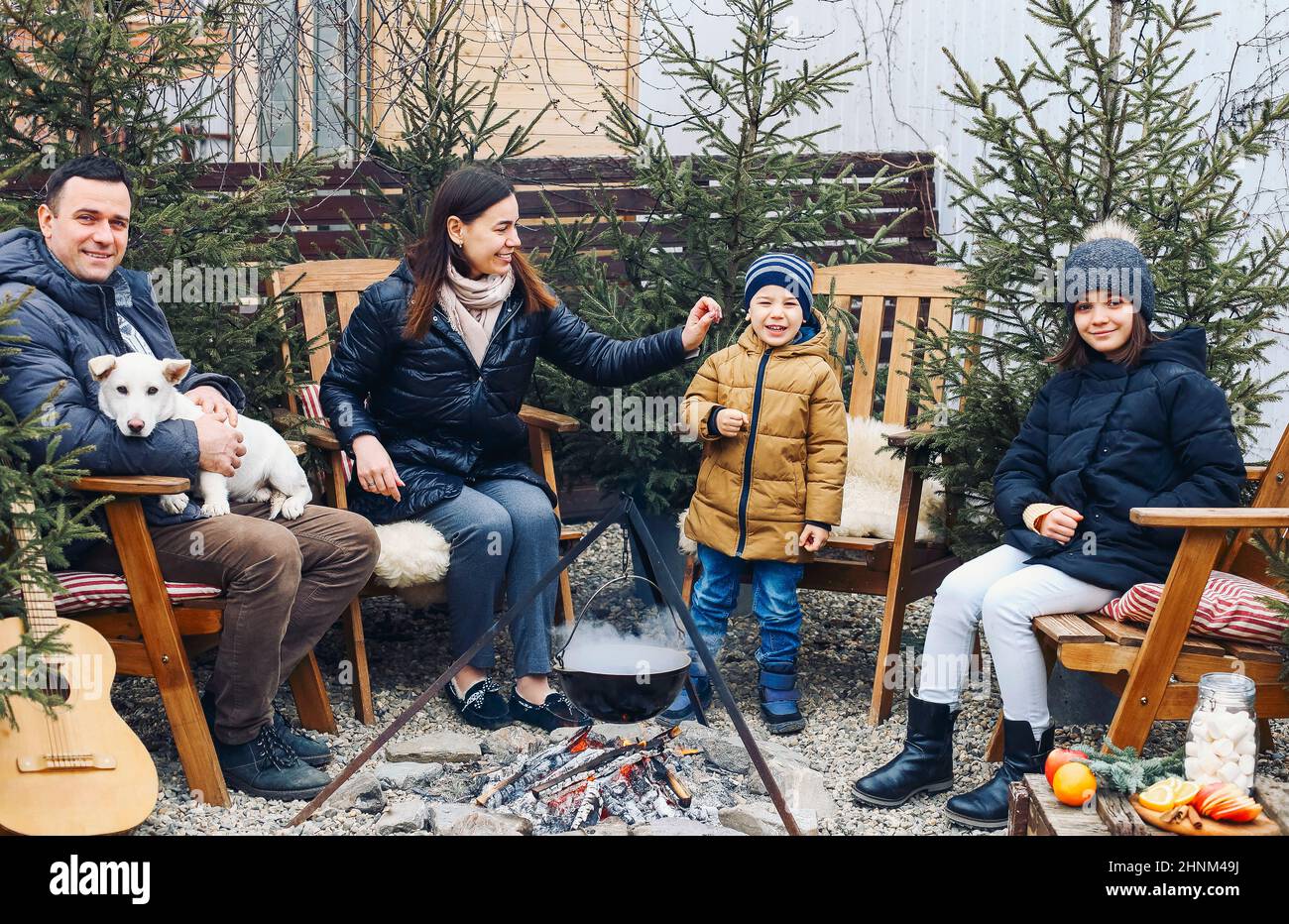 New Years gatherings by winter bonfire. Cozy portrait of happy family parents, two children and sweet dog cooking food in steaming pot over campfire w Stock Photo