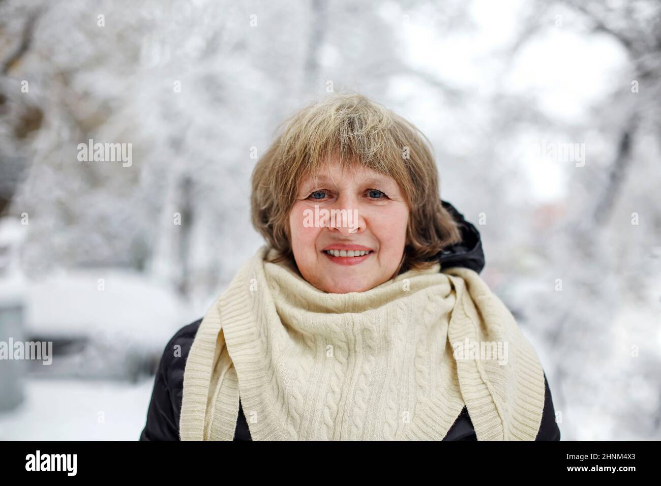 Portrait of happy pleased with frosty snowy weather elderly woman in warm clothes smiling at camera, standing in winter park outdoors during daily wal Stock Photo