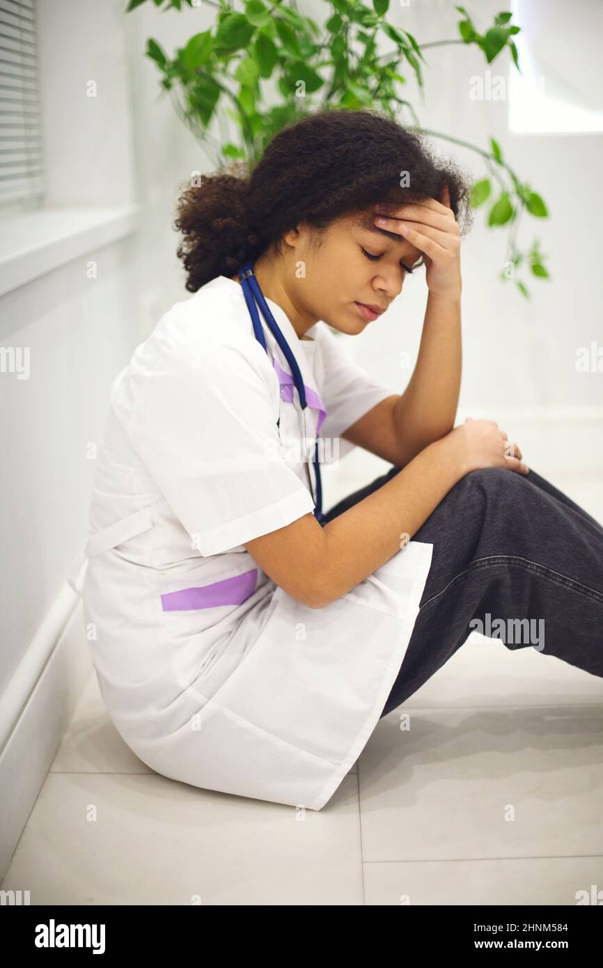 Sad depressed young african american nurse sitting on floor with frustrated face expression, feeling overwhelmed by workload in clinic, ethnic female Stock Photo
