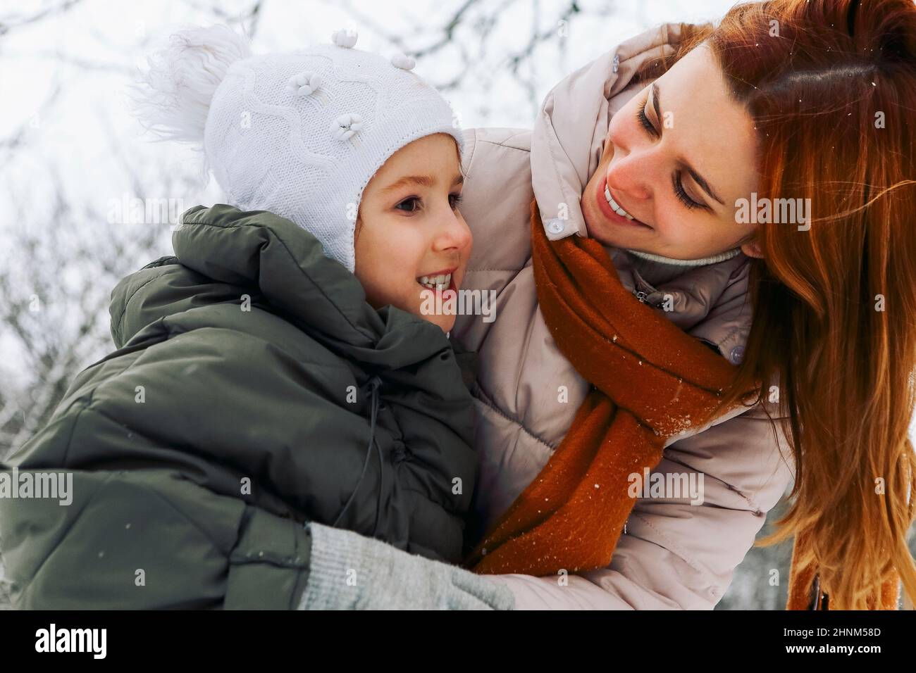 Lovely happy family mother and little girl daughter having fun on snowy weather outdoors, joyful playful mom with child hugging while spending time ac Stock Photo