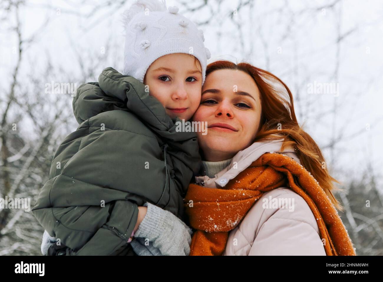 Lovely happy family mother and little girl daughter having fun on snowy weather outdoors, joyful playful mom with child hugging while spending time ac Stock Photo