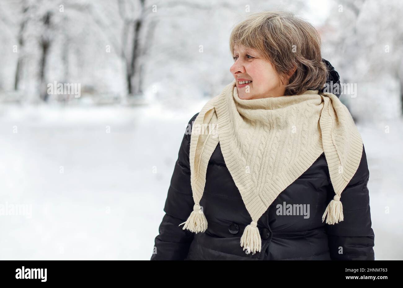 Portrait of happy pleased with frosty snowy weather elderly woman in warm clothes smiling at camera, standing in winter park outdoors during daily wal Stock Photo