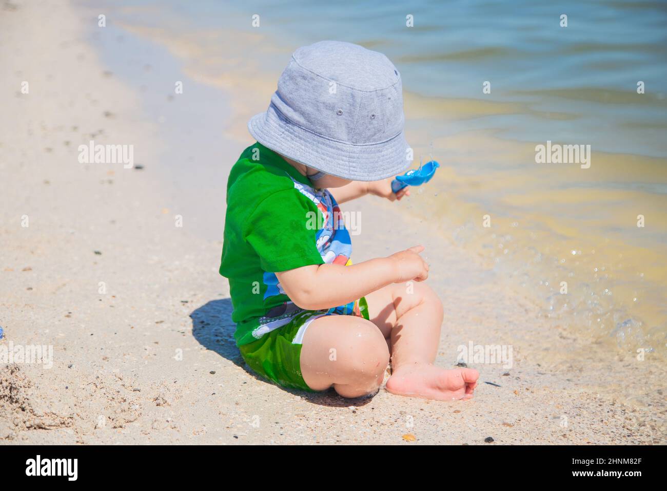 A little boy is sitting in a panama hat on the sand at the water's edge and playing with a shovel. Stock Photo
