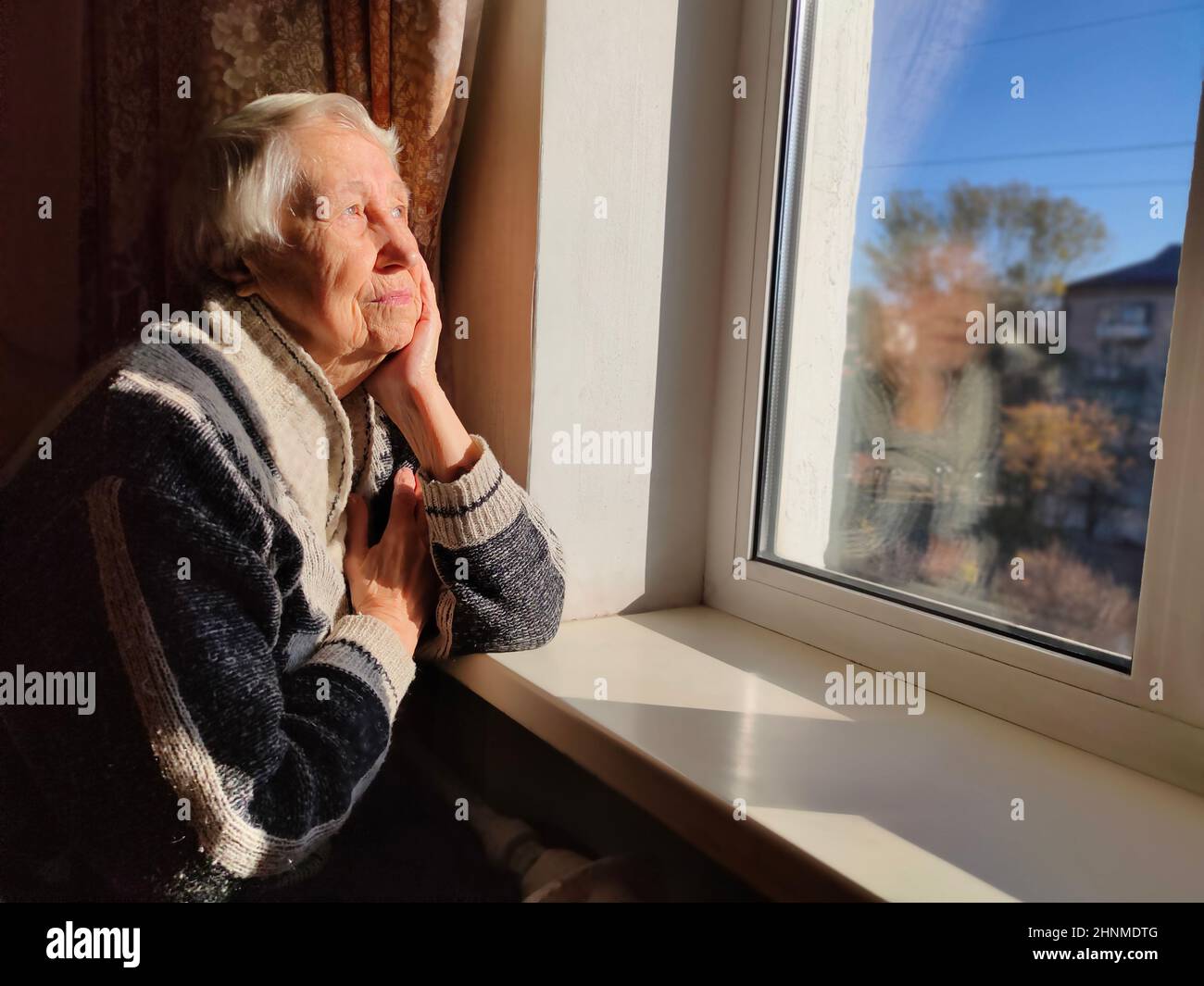 Old lonely woman sitting near the window in his house. Stock Photo