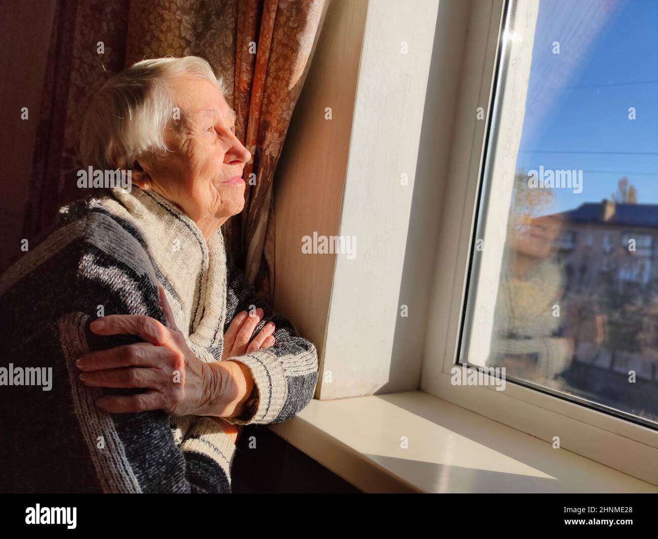 Old lonely woman sitting near the window in his house. Stock Photo