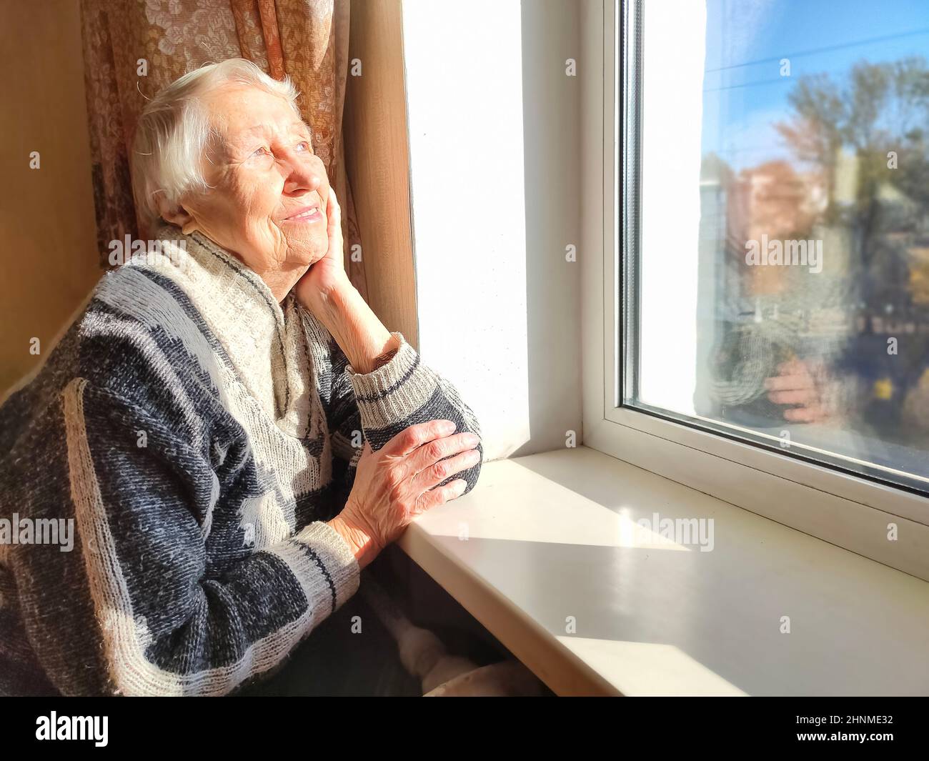Old lonely woman sitting near the window in his house. Stock Photo