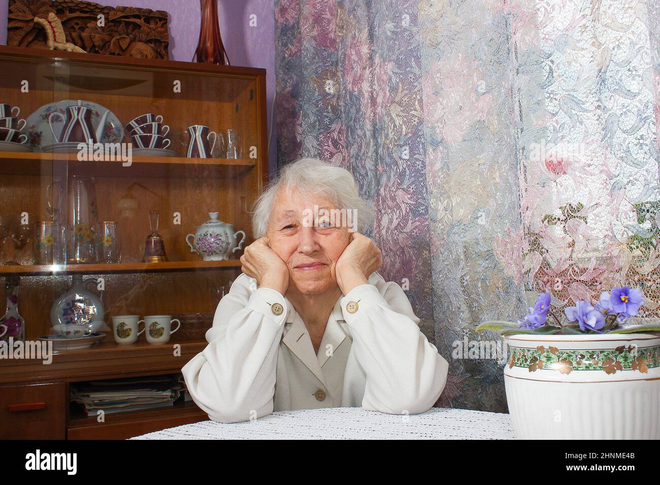 Old lonely woman sitting near the window in his house. Stock Photo