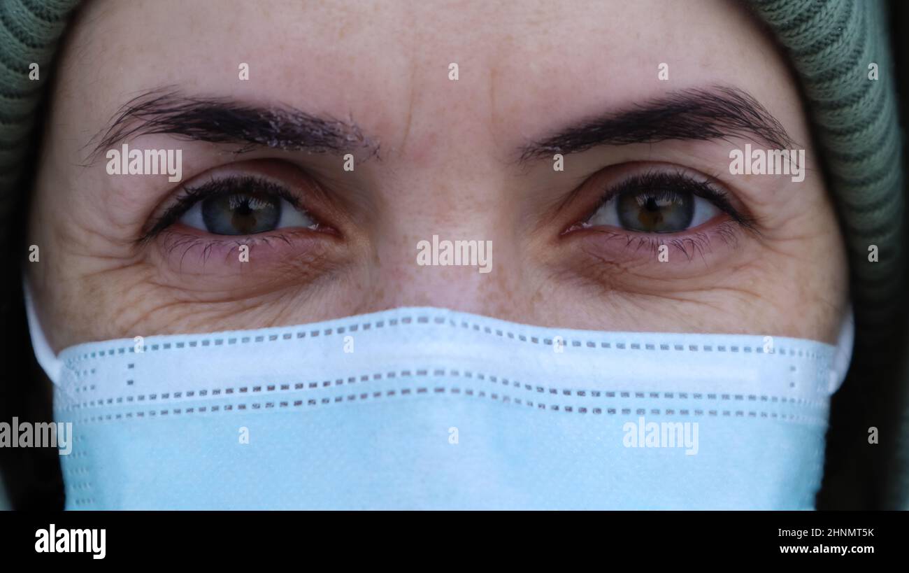 A beautiful young woman with a medical mask on her face is looking at you in the camera. Close-up portrait, fear in the eyes. Perfect eyes. Winter. Coronavirus protection. Health and safety concept. Stock Photo
