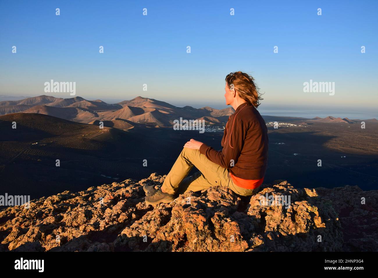 Sunrise in Lanzarote. A woman sitting on a mountain, overlooking the volcanic landscape with the small towns Yaiza and Uga. Canary Islands, Spain. Stock Photo