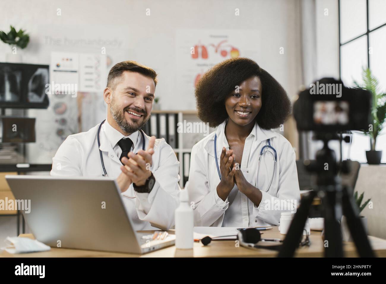 Caucasian male doctor and his african female nurse smiling and clapping in hands while recording broadcast at hospital office. Concept of people, telemedicine and technology. Stock Photo