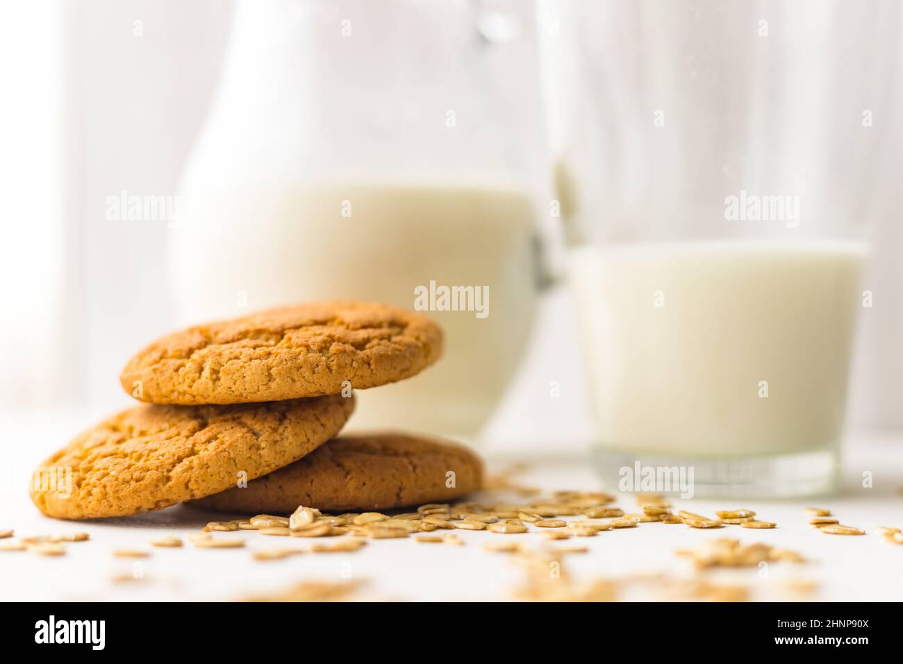 Pile oatmeal cookies and oat cereal flakes on white table on milk glass jug background Stock Photo