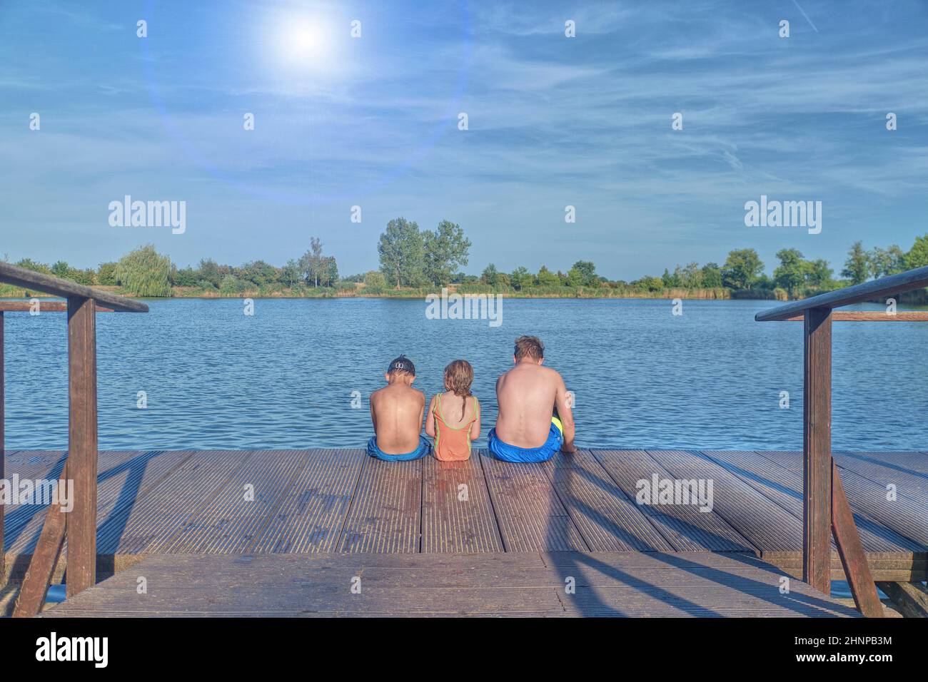 Children sitting on pier. Three children of different age - teenager boy, elementary age boy and preschool girl sitting on a wooden pier. Summer and childhood concept. Children on bench at the lake. Siblings sitting on wooden pier Stock Photo