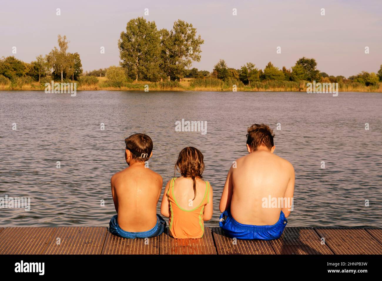 Children sitting on pier. Three children of different age - teenager boy, elementary age boy and preschool girl sitting on a wooden pier. Summer and childhood concept. Children on bench at the lake Stock Photo
