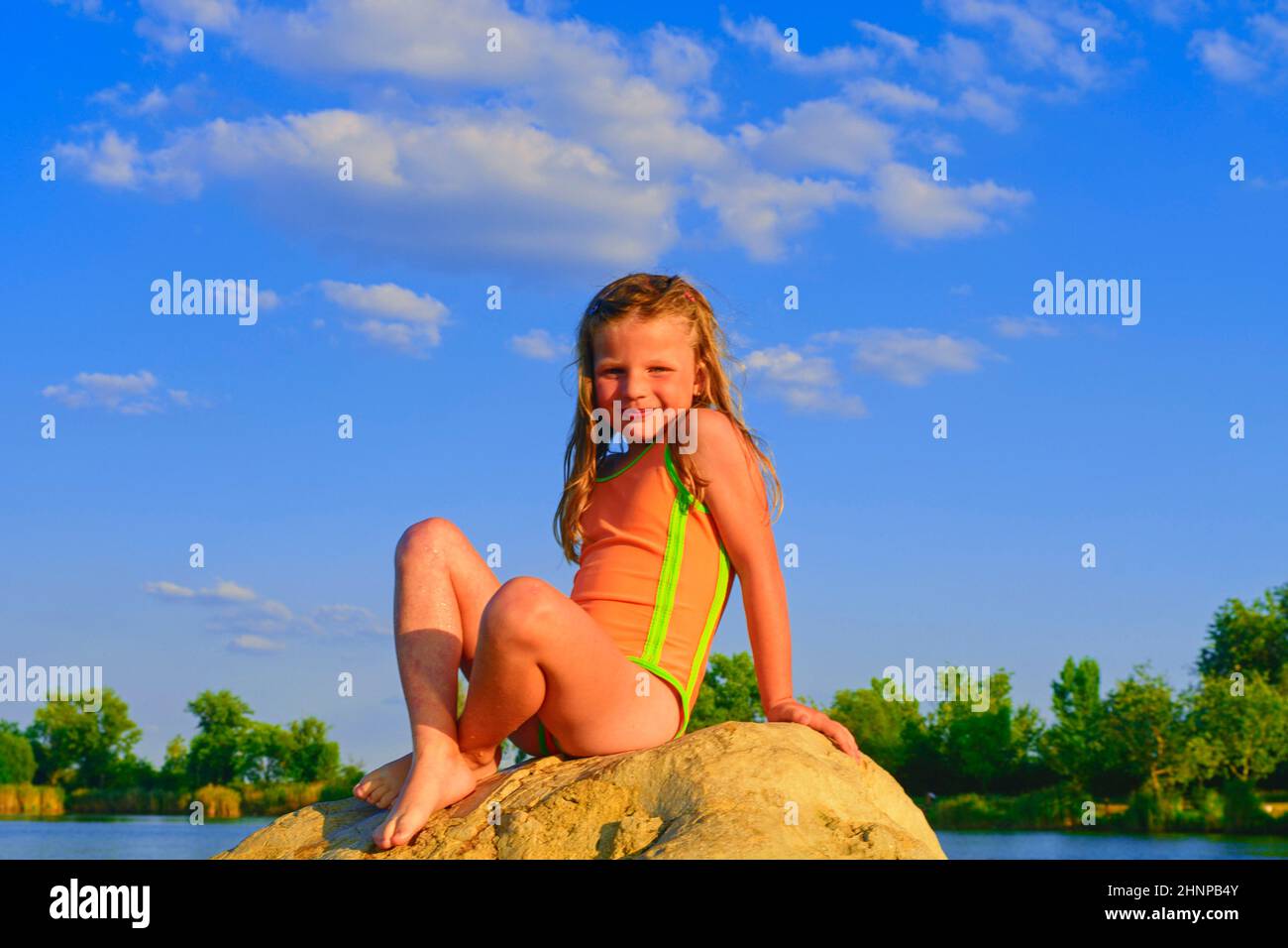 Beautiful girl sitting on a big rock. Little girl is wearing swimsuit. Summer and happy childhood concept. Copy space in bright blue sky Stock Photo