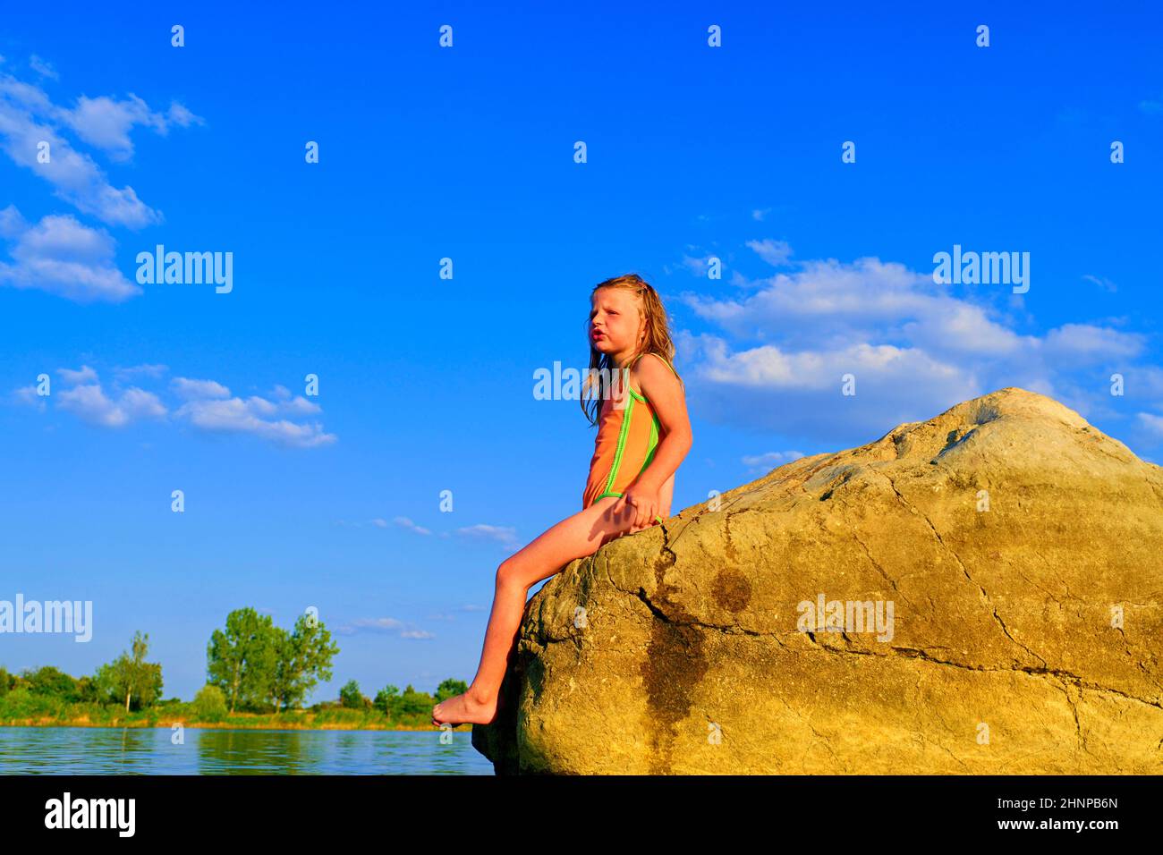 Beautiful girl sitting on a big rock. Little girl is wearing swimsuit. Summer and happy childhood concept. Copy space in bright blue sky Stock Photo