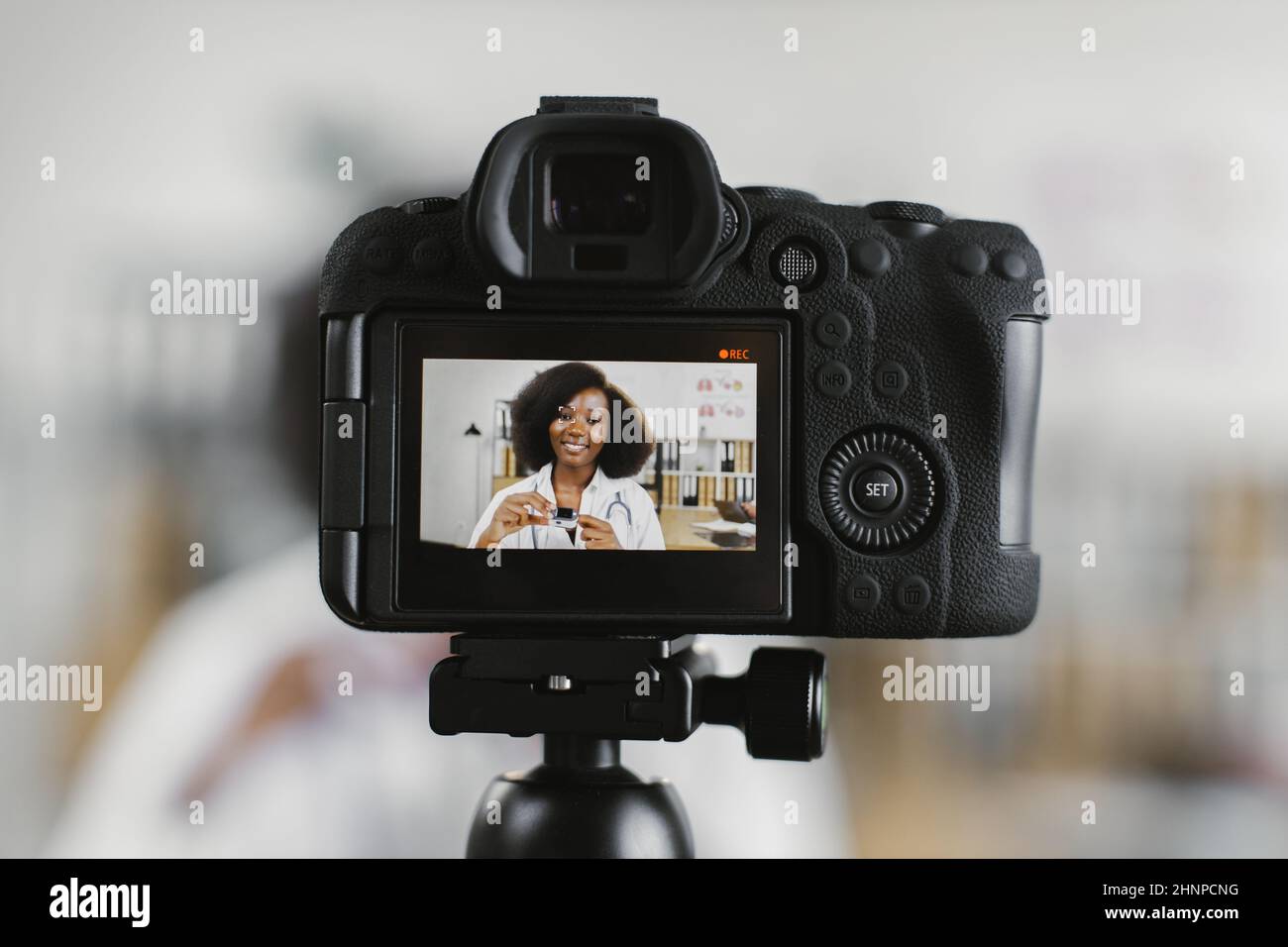 Video camera screen with pleasant african female doctor explaining how to use modern pulse oximeter. Competent therapist recording online tutorial at workplace. Stock Photo