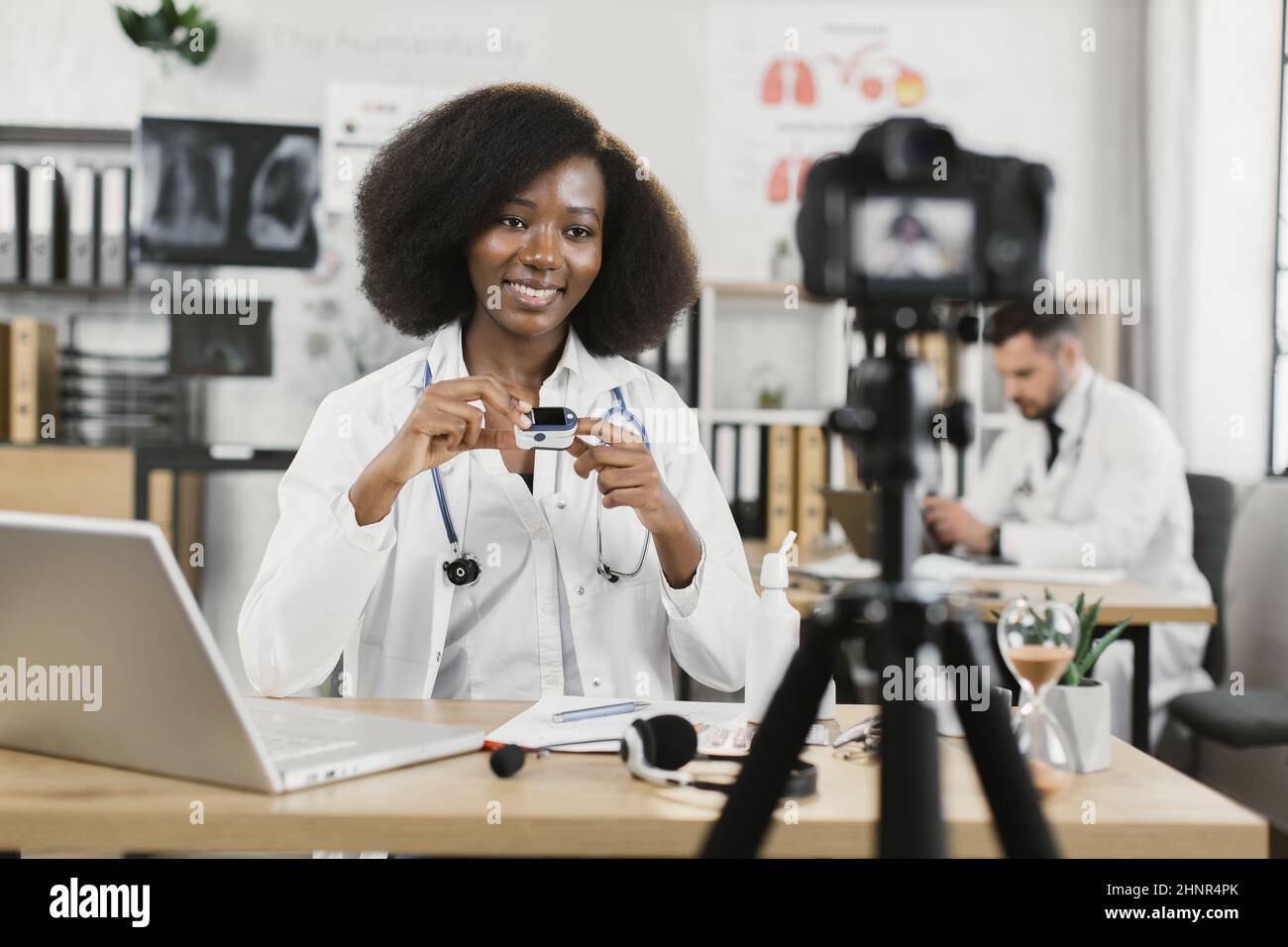 Positive african female doctor in lab coat recording video on camera and showing how to use pulse oximeter. Media channel with medical advice. Stock Photo