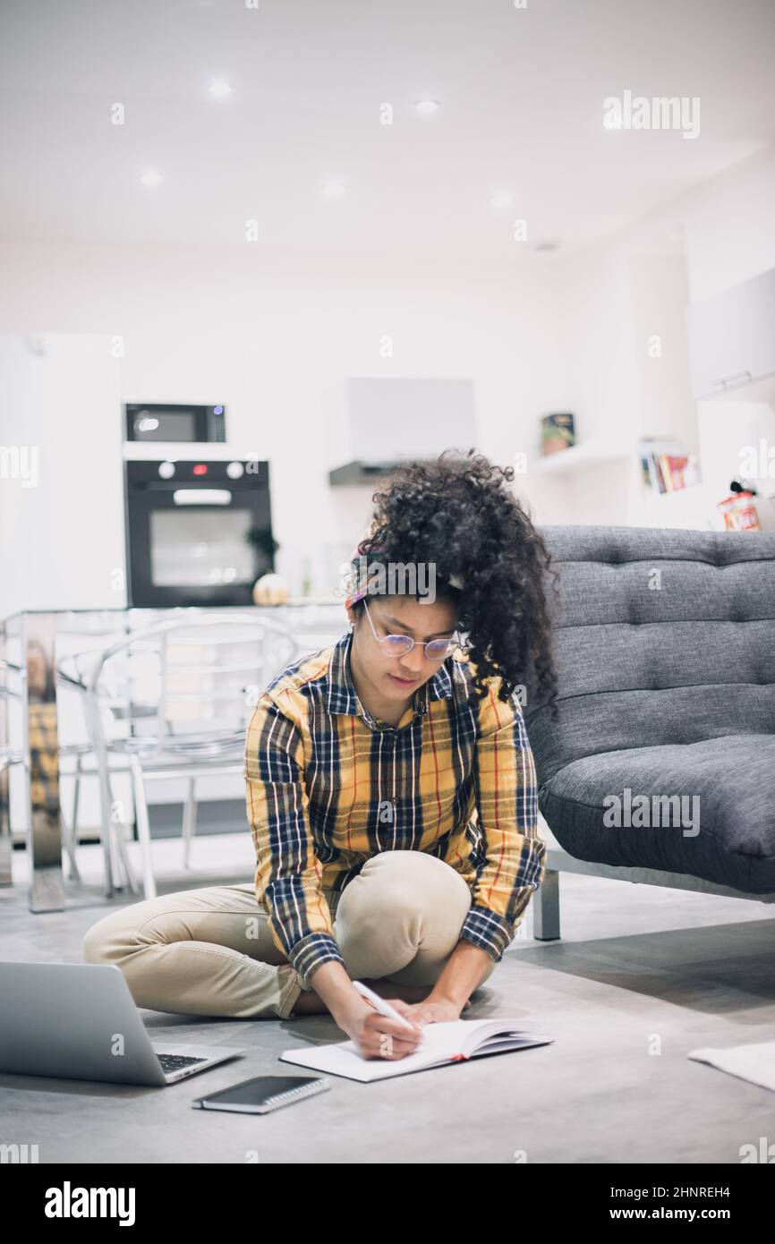 young Afro woman working from home sitting on floor with her laptop Stock Photo