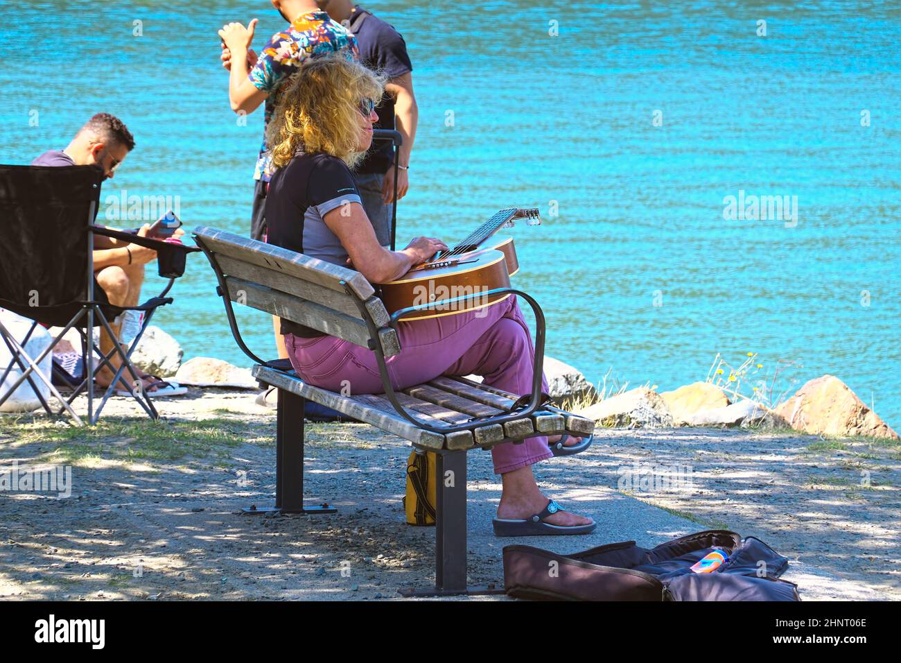 Blonde woman in sunglasses sitting with a guitar on a park bench beside a lake. Stock Photo