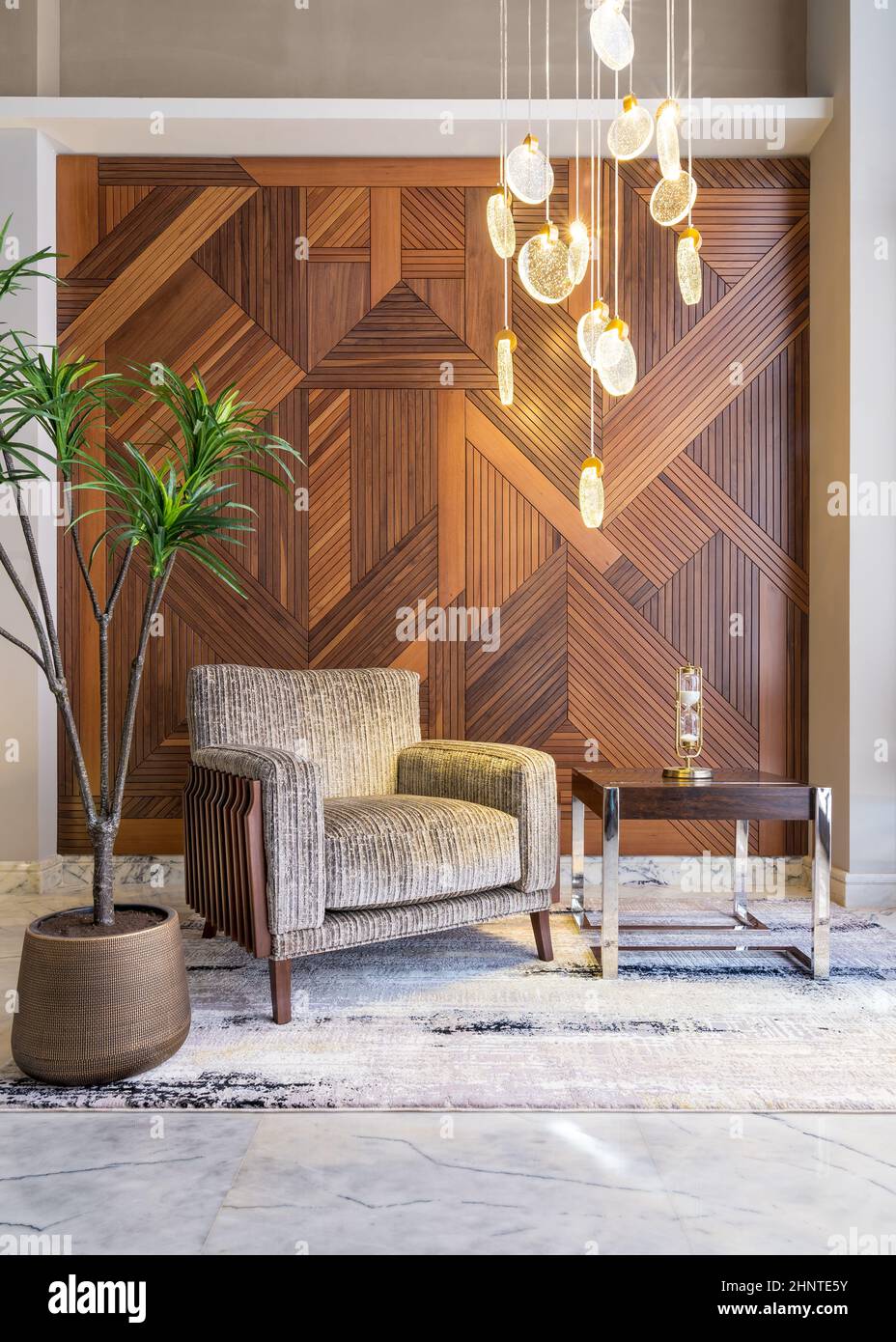 Modern beige armchair, small coffee table, planter, and modern tall glass chandelier, and decorated wood cladding wall Stock Photo