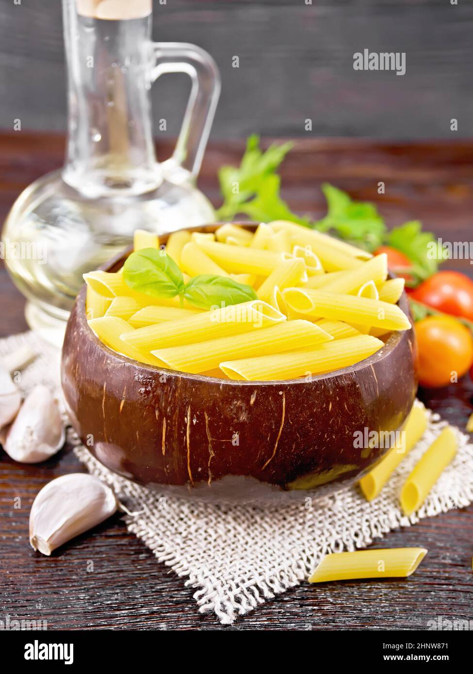 Wheat flour penne pasta in a bowl of coconut shells on sacking, tomatoes, garlic, vegetable oil in a decanter and parsley on wooden board background Stock Photo